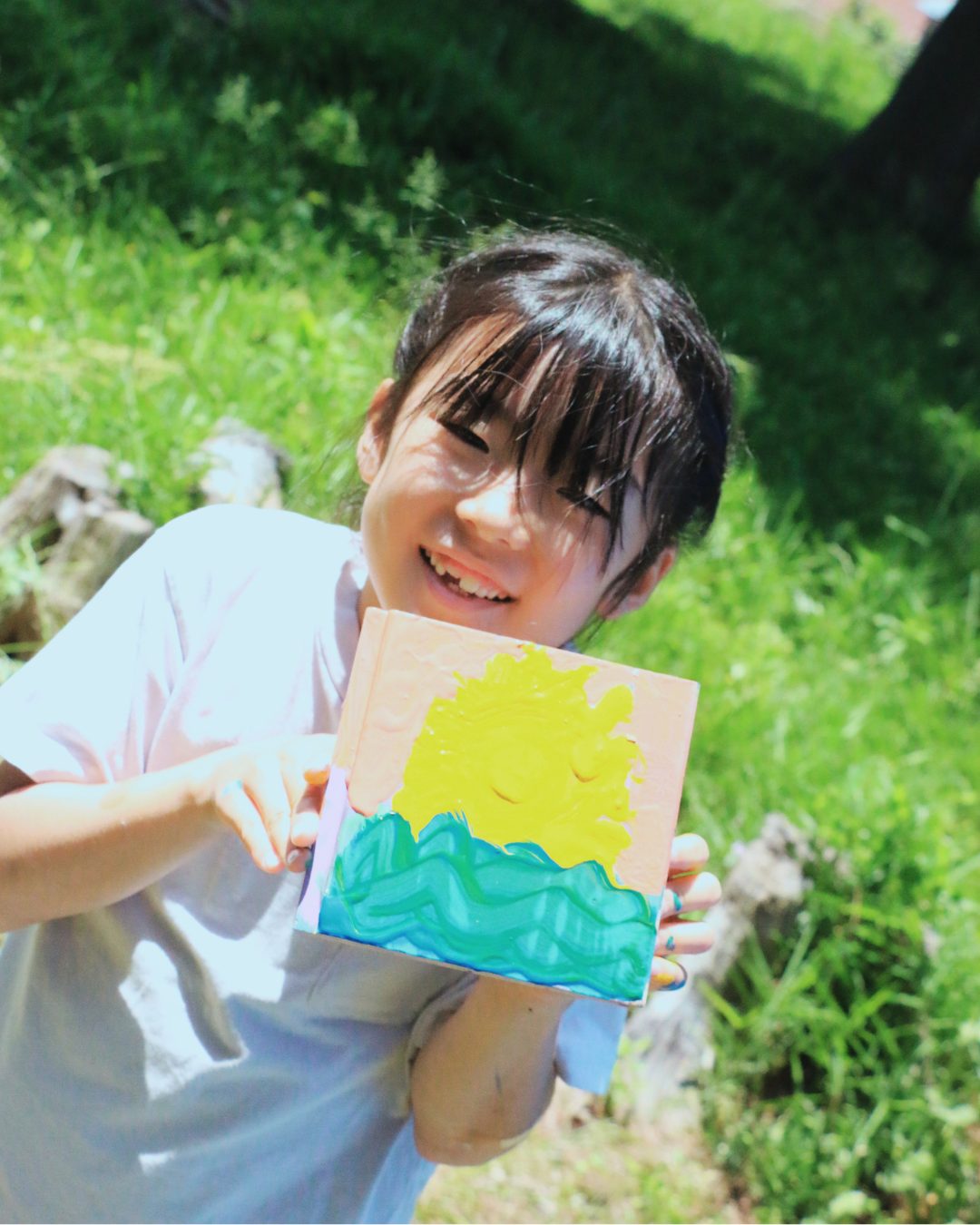 草の上で、太陽と海を描いた作品を掲げて笑う子ども