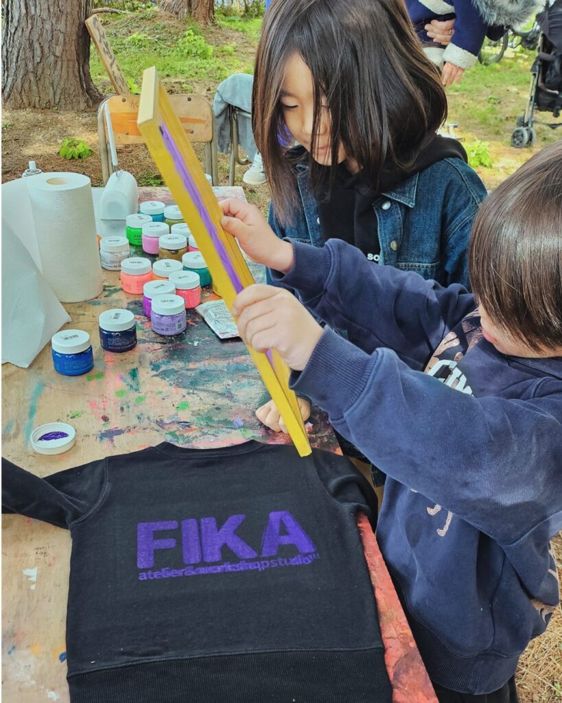 木陰の作業台で、シルクスクリーンの版を持ち上げる子どもたち。手前にプリントしたてのスウェット