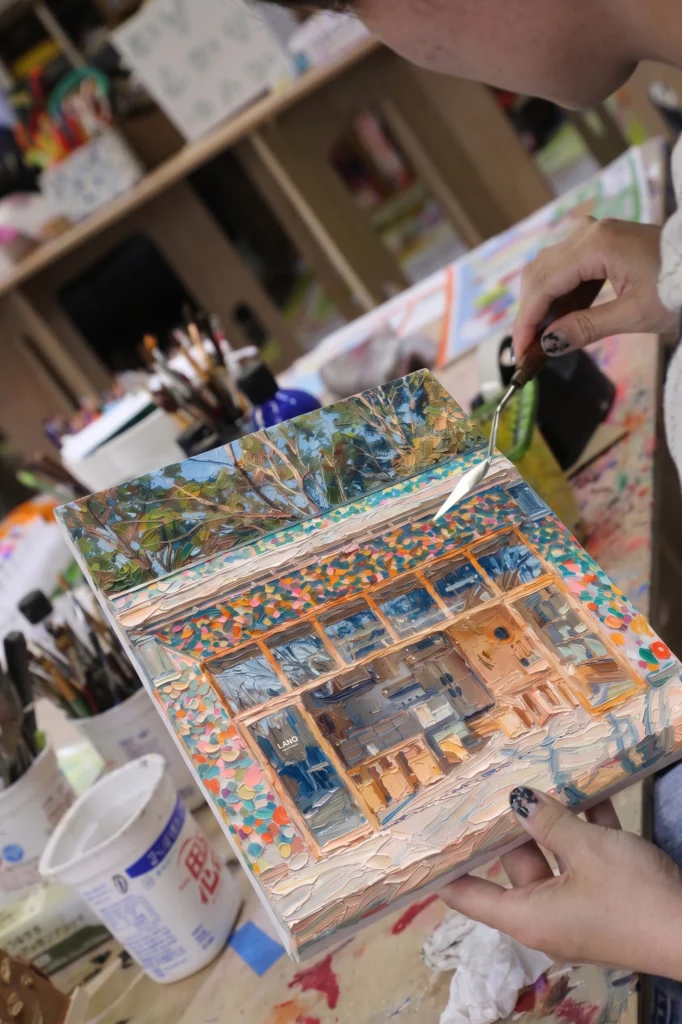 公園の木々と建物を描いたキャンバスに、パレットナイフで絵の具を厚くのせていく手元。建物の壁はオレンジ・水色・黄色の色とりどりのタイルのように塗られている