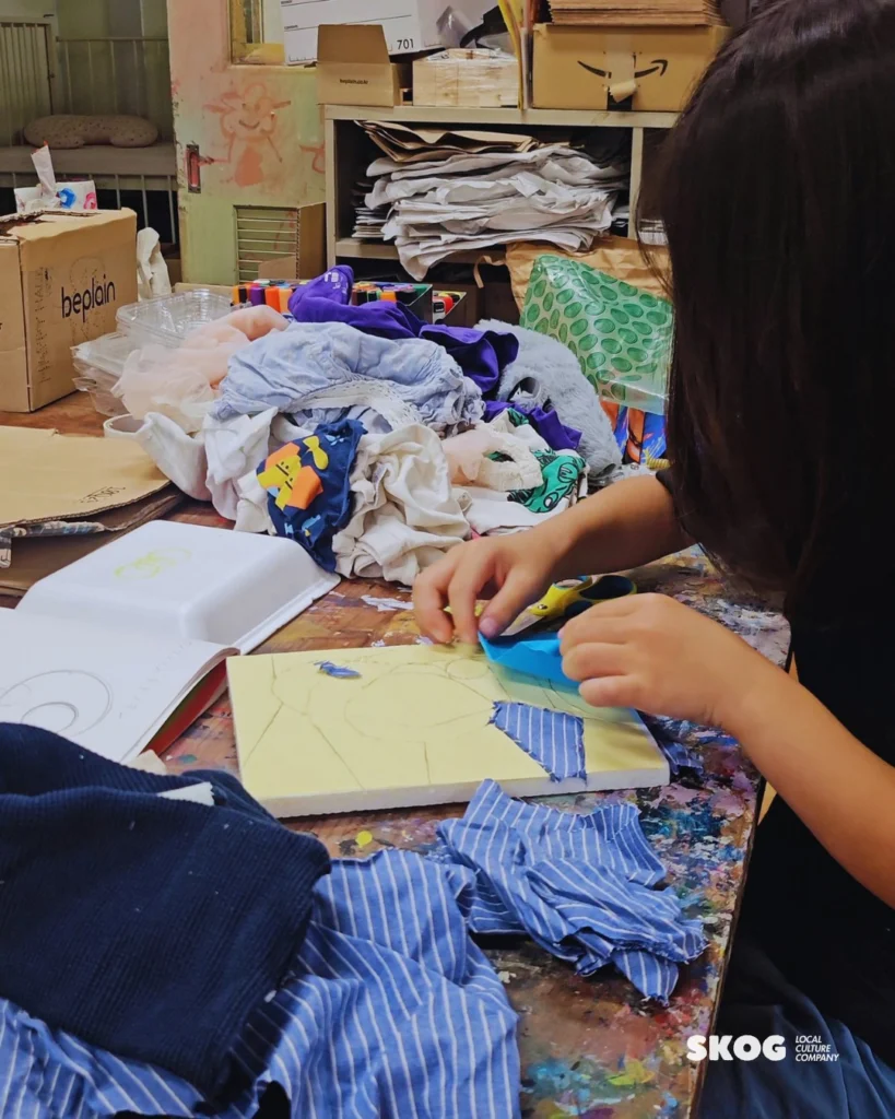 子どもが黄色い下絵のボードに、青い布の端切れを差し込んでいる。机の奥には服や布の端切れが積まれている